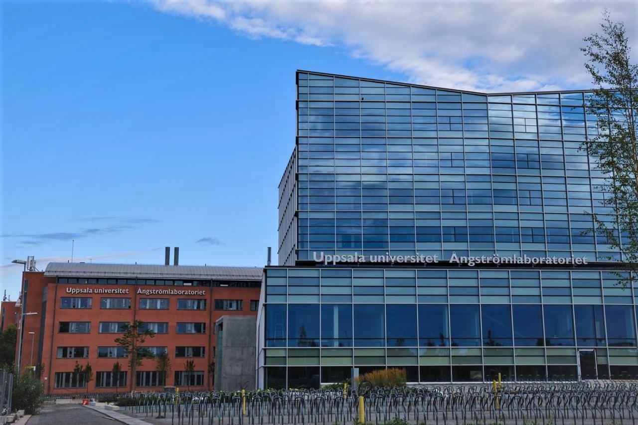 Modern glass building of Uppsala University’s Ångström Laboratory with bike racks in front under a blue sky.